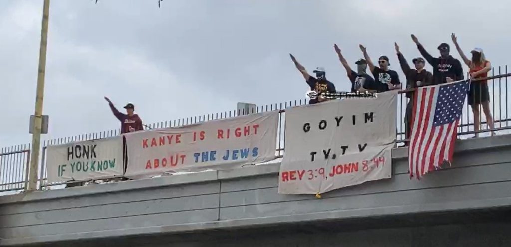 Group of protesters making a Nazi salute while holding an American flag and signs over a highway overpass which read, "Honk if you know," "Kanye is right about the Jews," and "GoyimTV dot TV Rev 3:9 John 8:44."